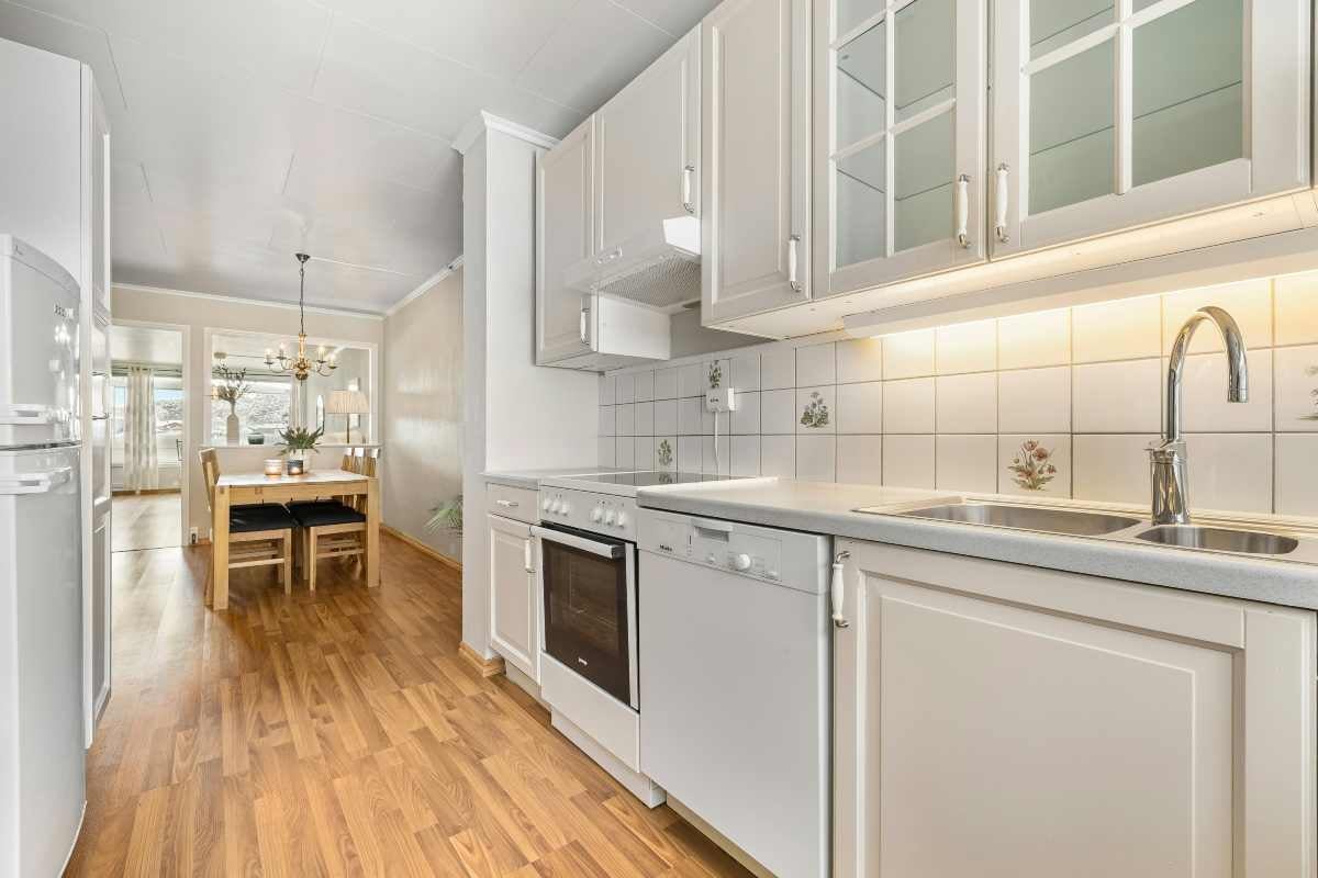 Bucătărie luminoasă cu dulapuri albe, electrocasnice și pardoseală din lemn, în fundal un loc de luat masa cu masă și scaune din lemn.