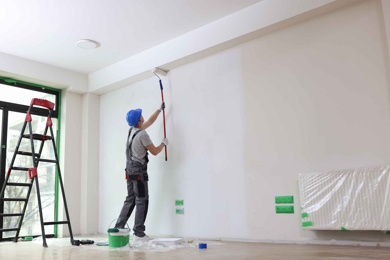 Un zugrav vopsește un perete cu un trafalet într-o cameră luminoasă, incluzând o scară și o găleată cu vopsea.