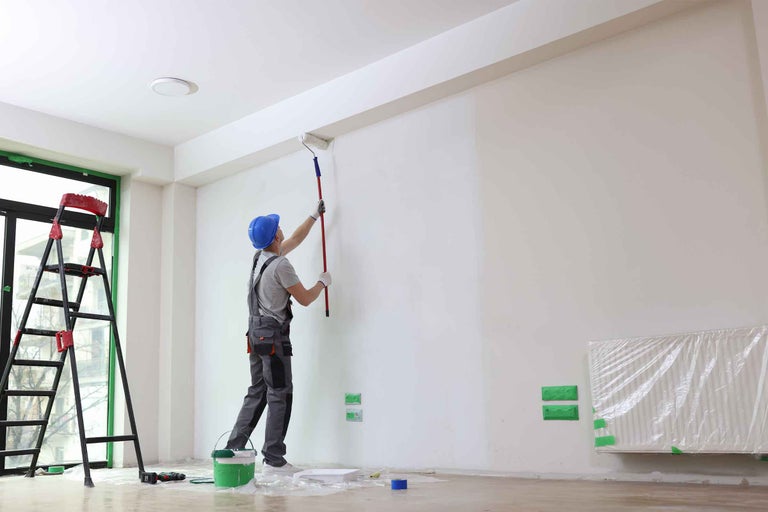 Un zugrav vopsește un perete cu un trafalet într-o cameră luminoasă, incluzând o scară și o găleată cu vopsea.