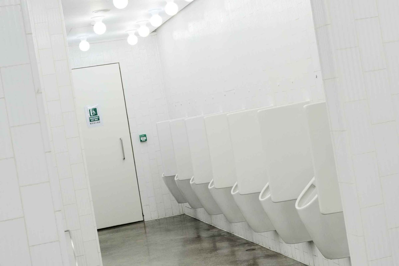 Toaletă publică pentru bărbați cu un rând de pisoare montate pe un perete cu faianță, spațiu luminos cu ieșire de urgență.