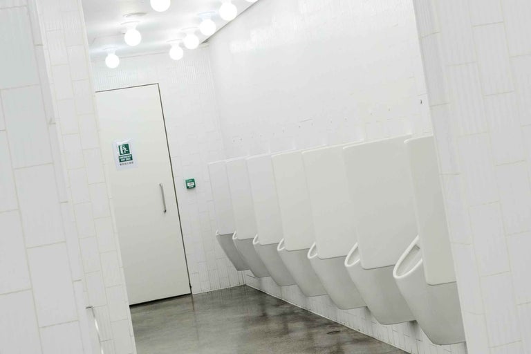 Toaletă publică pentru bărbați cu un rând de pisoare montate pe un perete cu faianță, spațiu luminos cu ieșire de urgență.