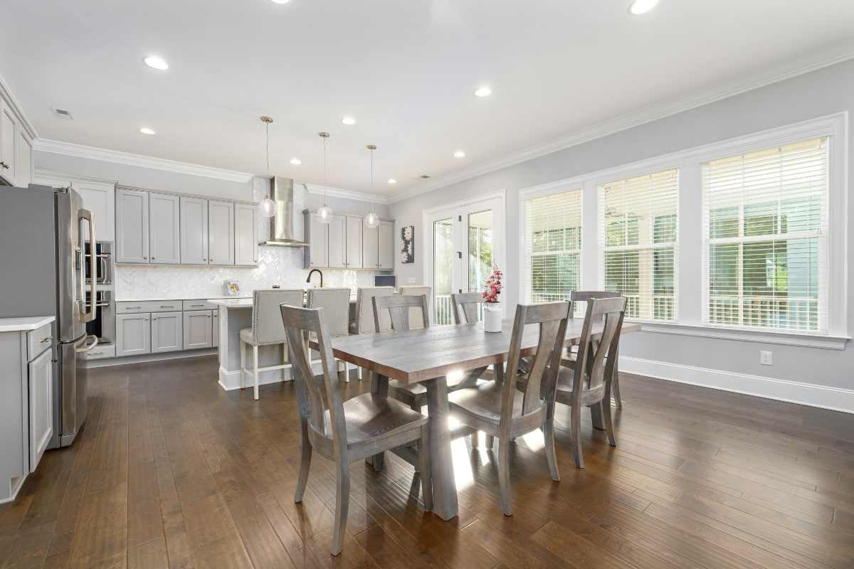 Bucătărie și zonă de dining modernă și luminoasă, cu o masă mare din lemn, scaune din lemn asortate, dulapuri gri deschis și podea din lemn închis.