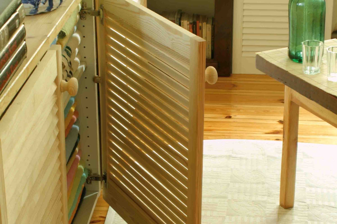 Ușă lamelară deschisă din lemn de pin la un dulap, într-o încăpere luminoasă cu pardoseală din lemn și covor.