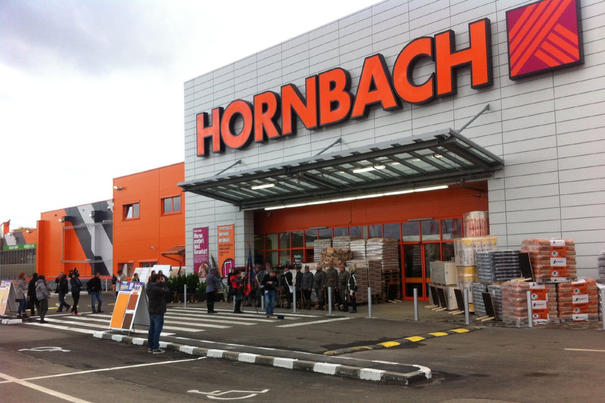 Vedere exterioară a unui magazin de bricolaj Hornbach cu clienți și mărfuri în fața intrării