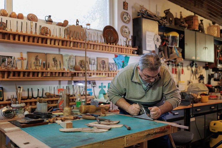 Un bărbat rindeluiește lemn într-un atelier cu unelte și rafturi.