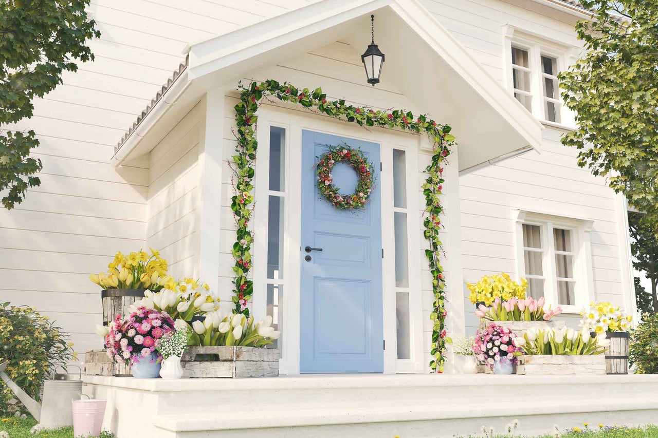 Fațadă luminoasă de casă cu intrare, decorată cu flori de primăvară, o coroniță de ușă și ghirlande de flori.