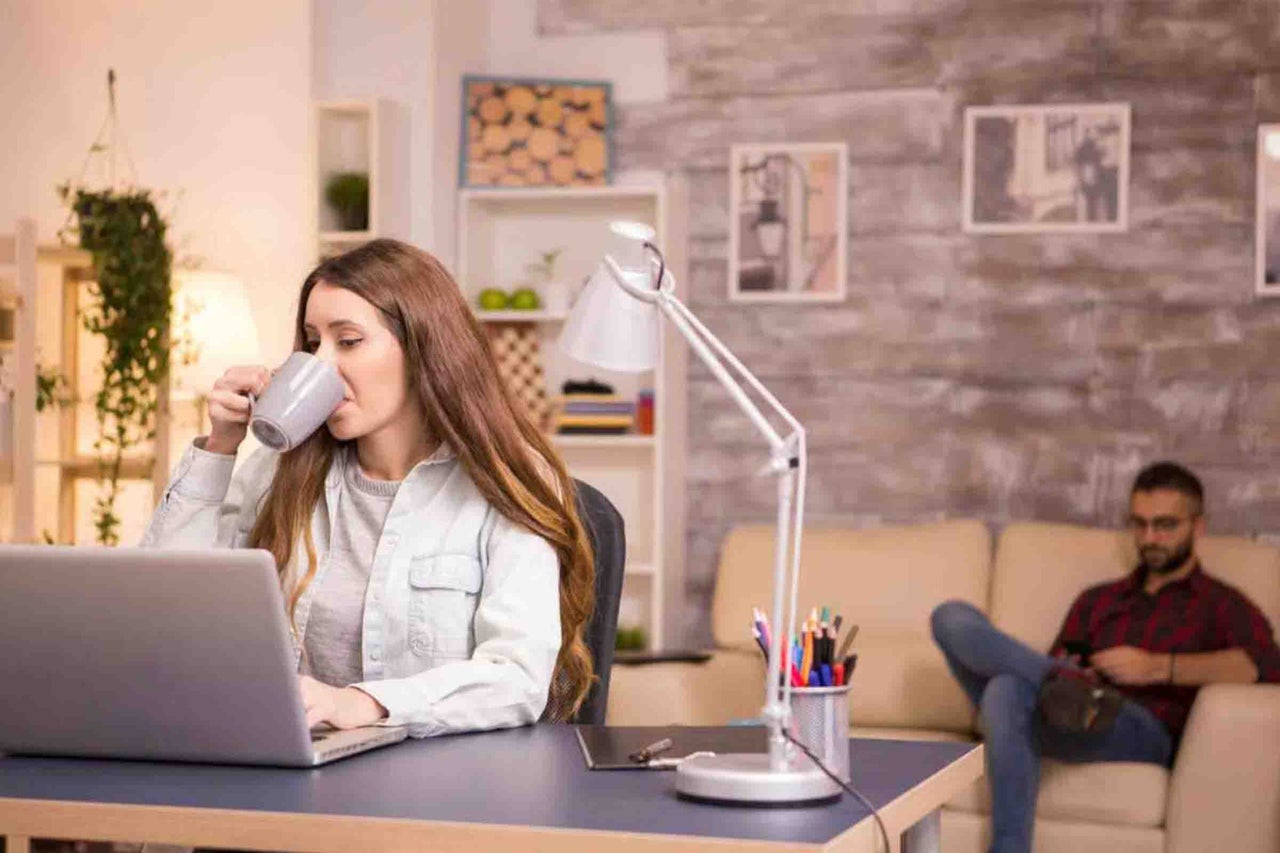 O femeie lucrează pe un laptop la un birou cu o lampă de birou albă, în timp ce un bărbat stă pe o canapea în fundalul unui birou de acasă.
