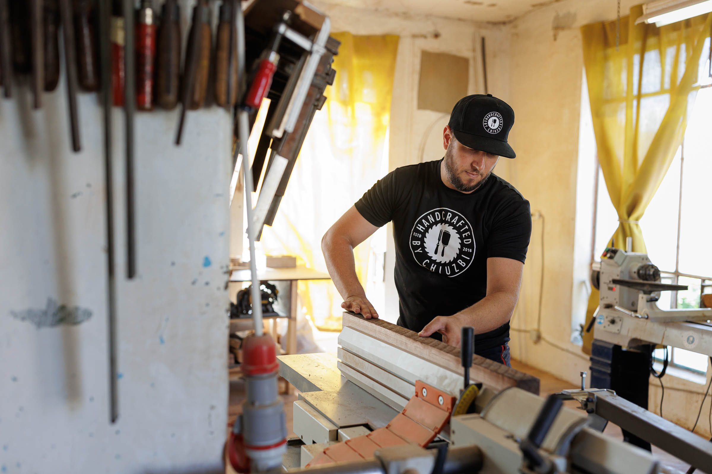 Un bărbat lucrează cu un ferăstrău circular de masă într-un atelier. Unelte sunt fixate pe perete.