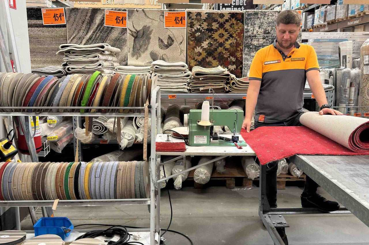 Un angajat Hornbach taie și prelucrează un covor într-un magazin de bricolaj Hornbach. Pe fundal sunt expuse diverse covoare și borduri.