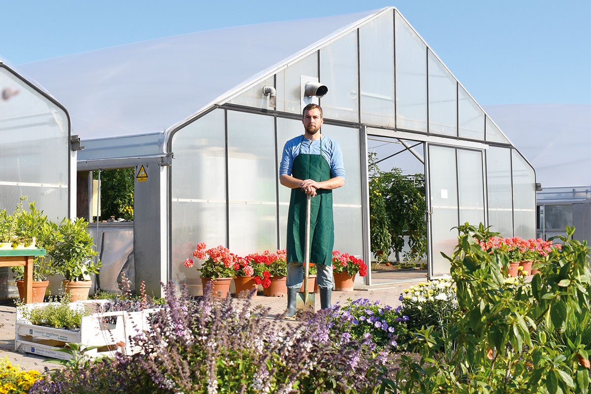 Un grădinar stă în fața unei sere cu flori și plante.