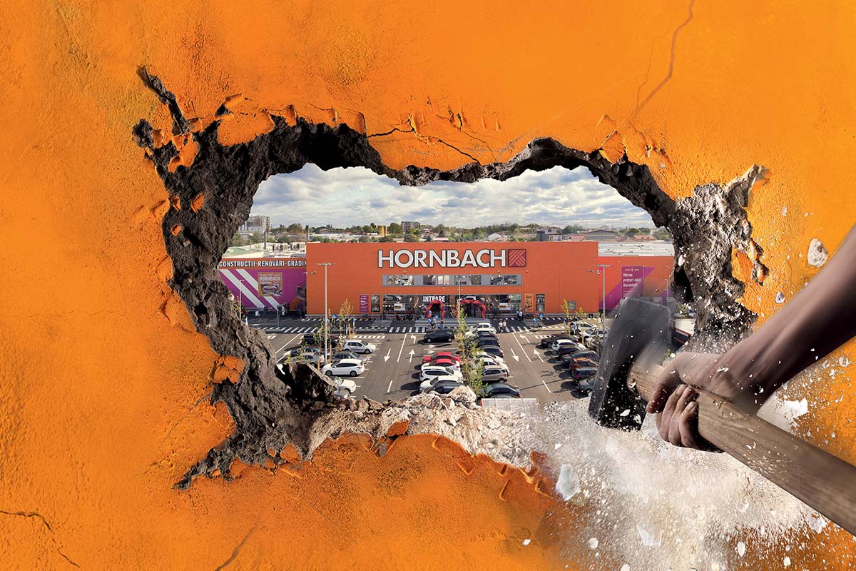 Vedere printr-o gaură într-un perete spre un magazin de bricolaj Hornbach cu parcare. O persoană lovește peretele cu un ciocan.