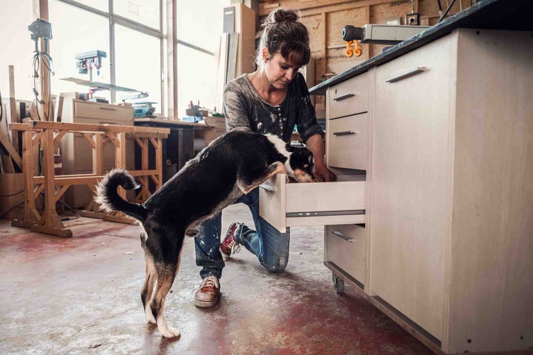Femeie îngenuncheată cu un câine în fața unui dulap de atelier cu sertare
