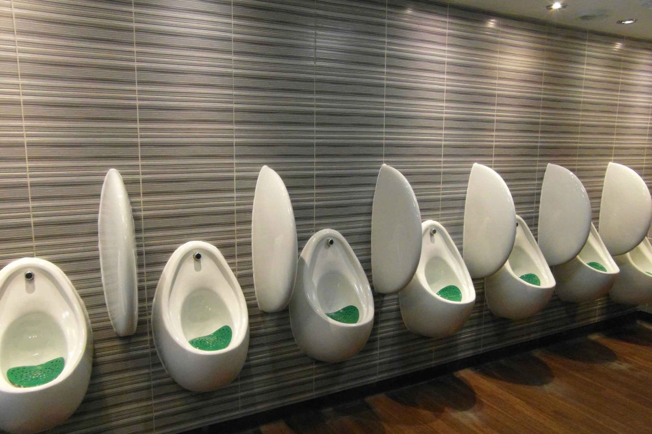 Toaletă publică modernă cu un rând de pisoare suspendate din ceramică cu separatoare în fața unui perete cu plăci ceramice cu dungi.