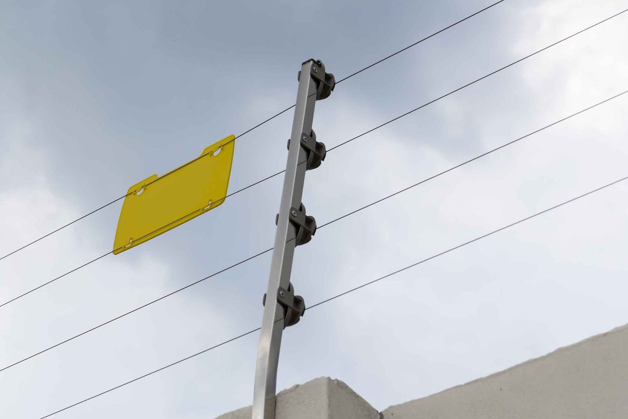 Stâlp metalic pentru gard electric cu izolatori, fire și plăcuță de avertizare montat pe un zid.