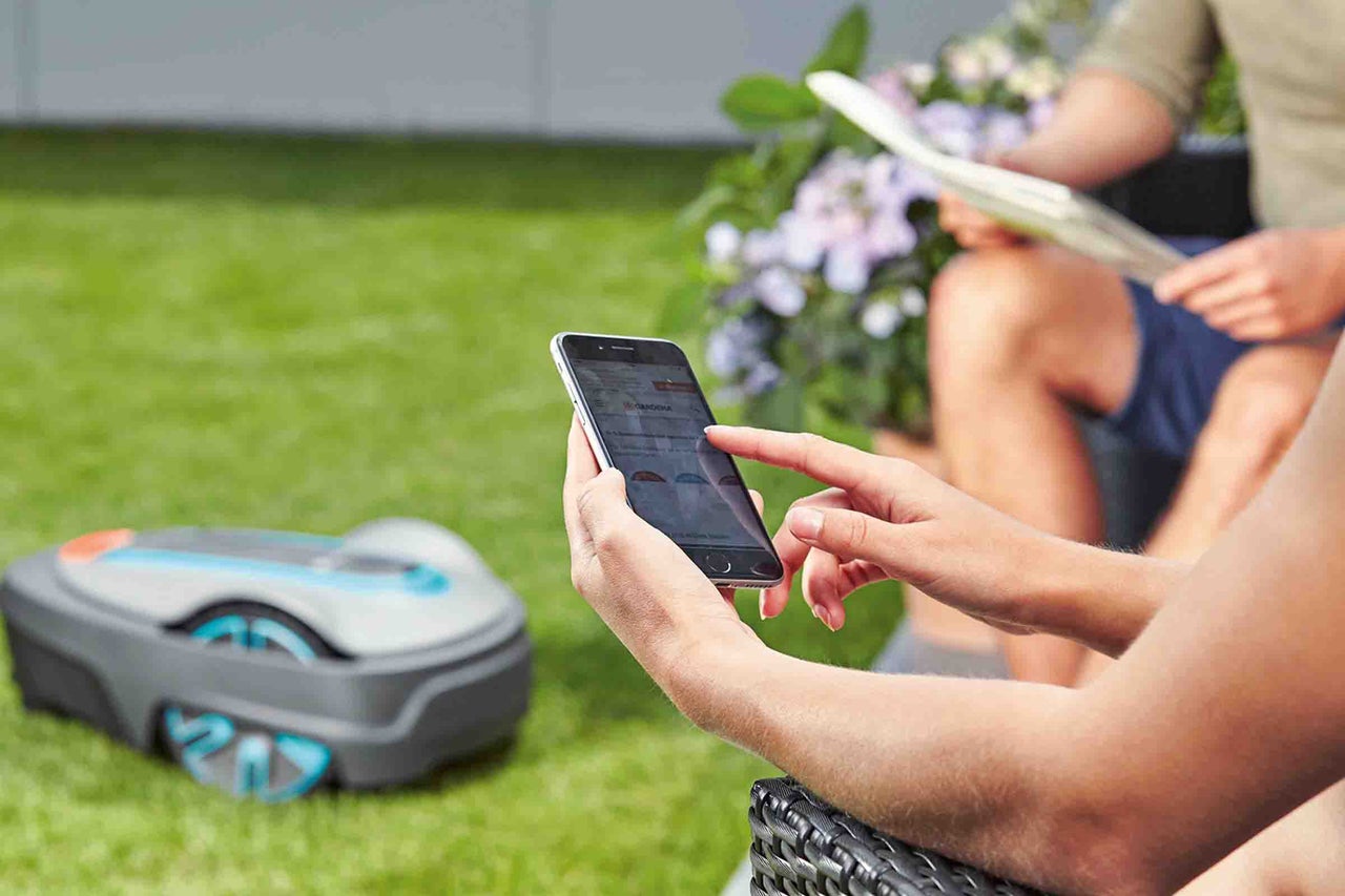 O persoană operează o aplicație pe un smartphone lângă o mașină de tuns iarba robotizată pe un gazon.