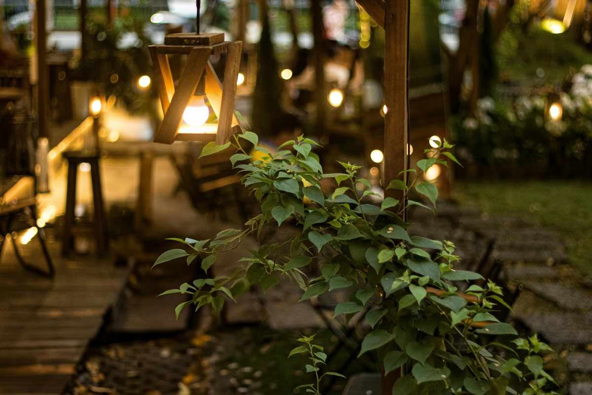 Lanternă de grădină suspendată din lemn cu lumină caldă într-o grădină iluminată cu plante verzi și design rustic.
