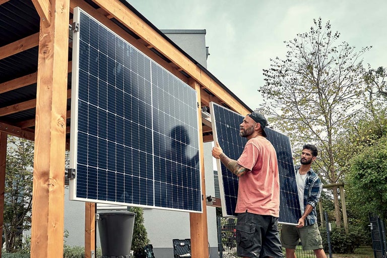 Doi bărbați montează un panou fotovoltaic pe structura de lemn a unui adăpost auto din grădină.