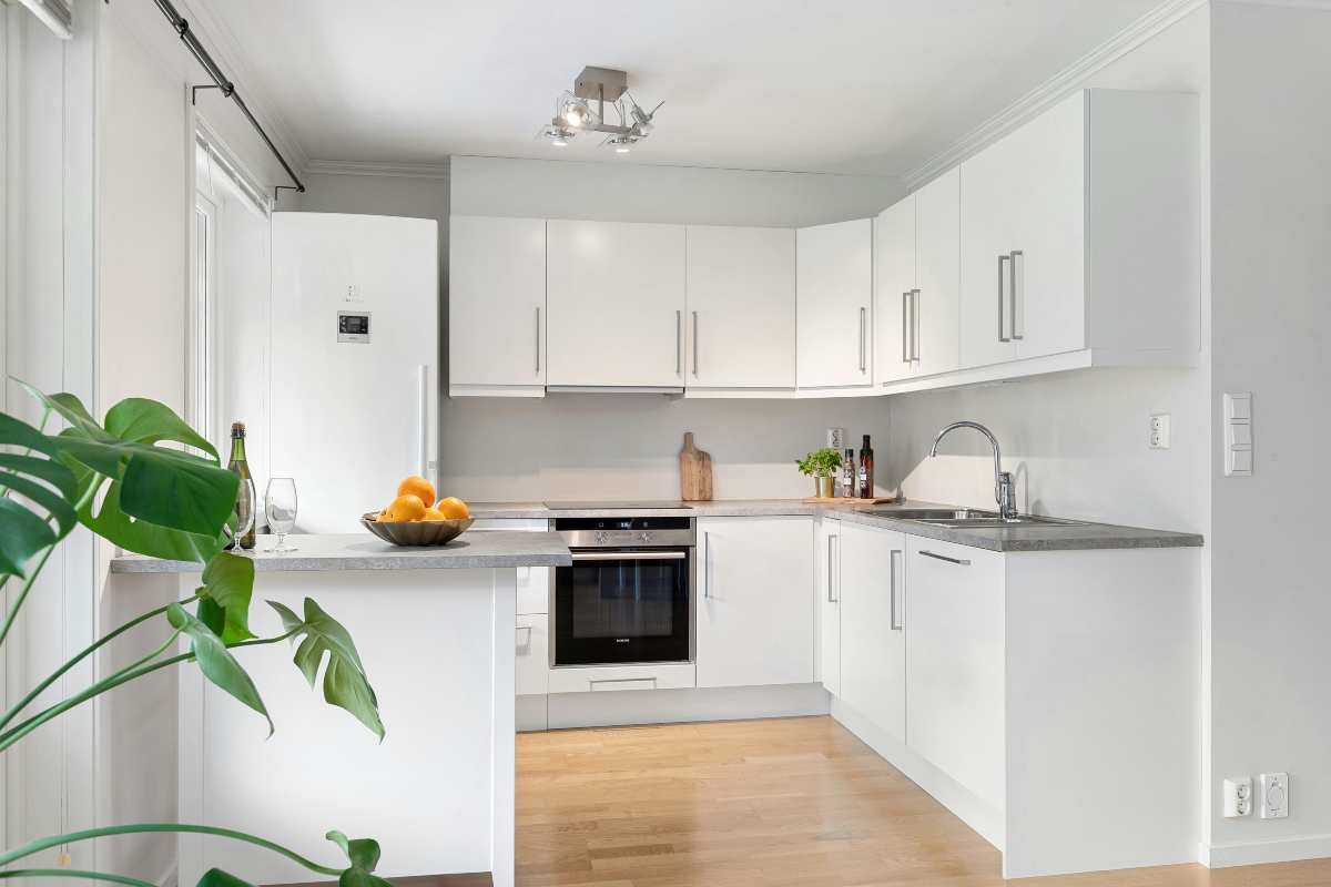 Bucătărie modernă în formă de L, cu fronturi albe, blat gri, cuptor încorporat și podea din lemn.