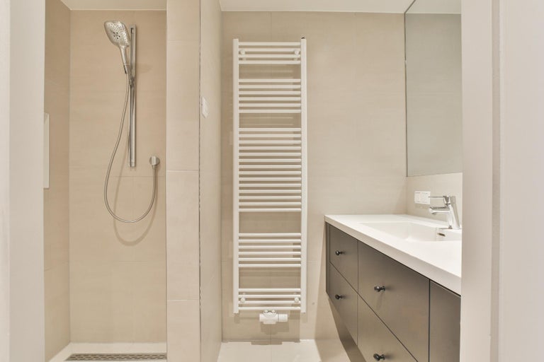Baie modernă și luminoasă cu duș de tip walk-in, radiator port-prosop alb și mască de chiuvetă închisă la culoare cu lavoar alb.