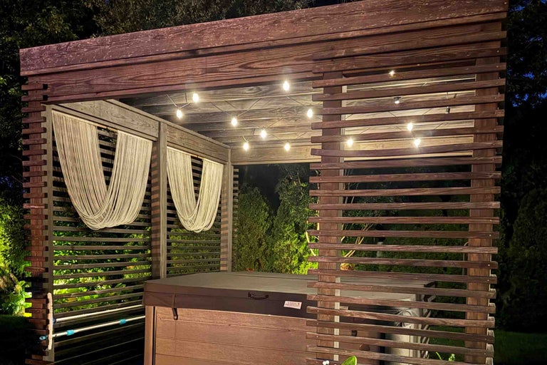 Un pavilion de grădină din lemn cu o cadă cu hidromasaj acoperită în interior, iluminat de o ghirlandă de lumini noaptea.