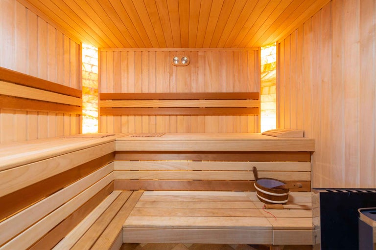 Sauna interioara moderna din lemn deschis la culoare cu banci de colt, iluminat din pietre de sare pe peretii laterali si galeata de sauna.