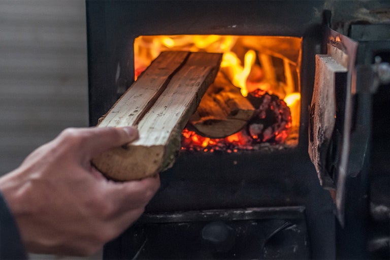 O mână pune lemne într-un șemineu pe lemne în care arde deja un foc.