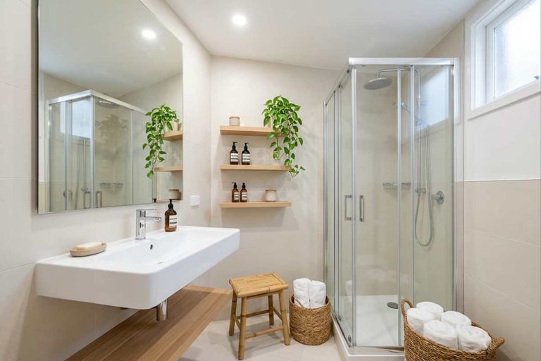 Baie modernă și luminoasă cu lavoar suspendat, oglindă și cabină de duș din sticlă. Cu rafturi suspendate din lemn, scaun din bambus și coșuri.