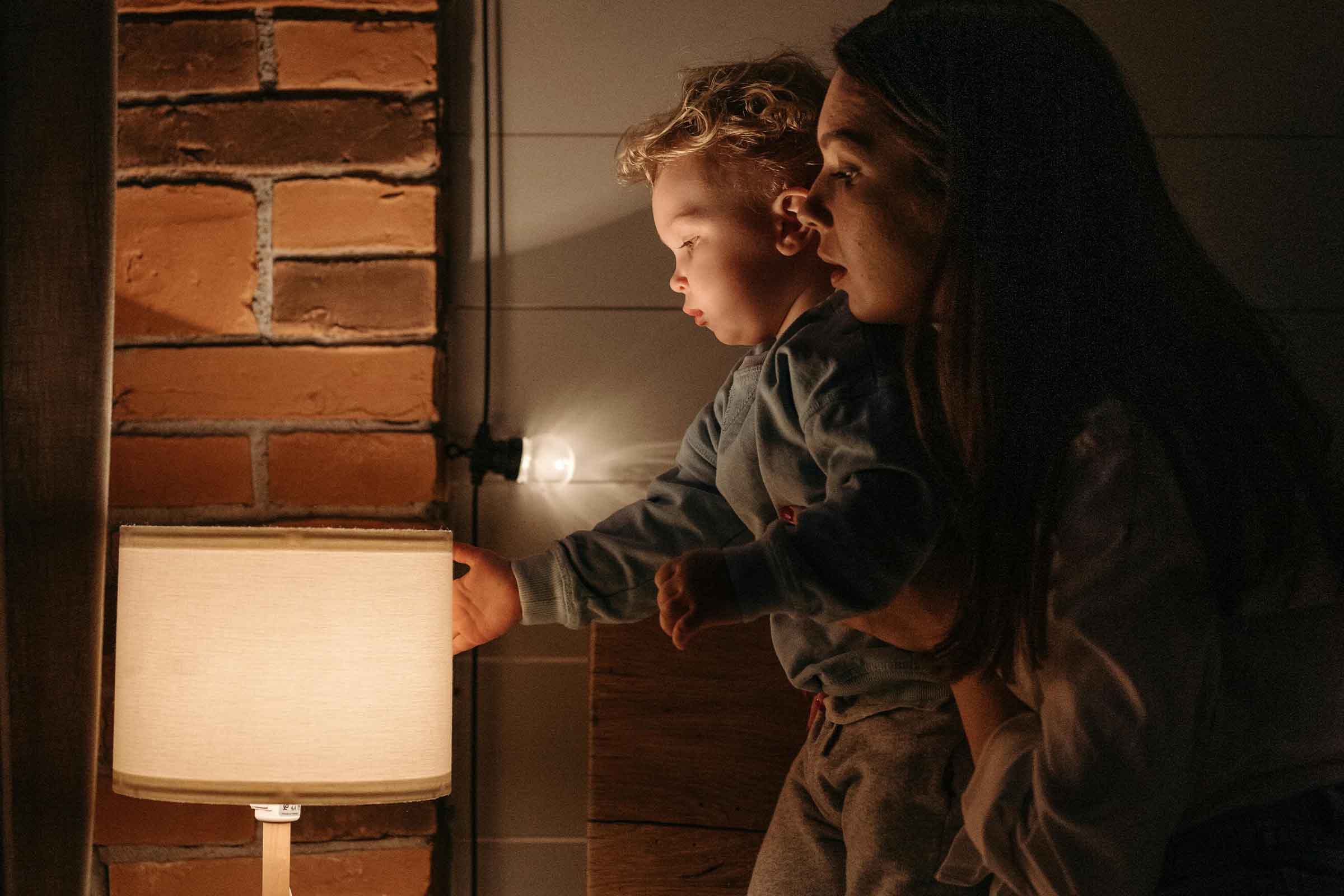 Mamă ținând un copil mic lângă o lampă aprinsă în fața unui zid de cărămidă
