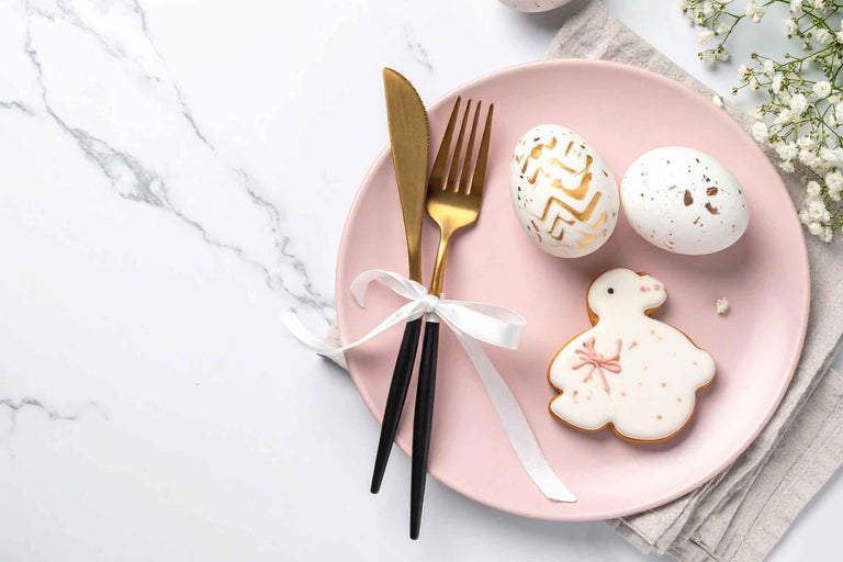 Decorațiune de masă de Paște cu o farfurie roz, tacâmuri aurii cu mânere negre, ouă de Paște decorate și un biscuite în formă de iepuraș.