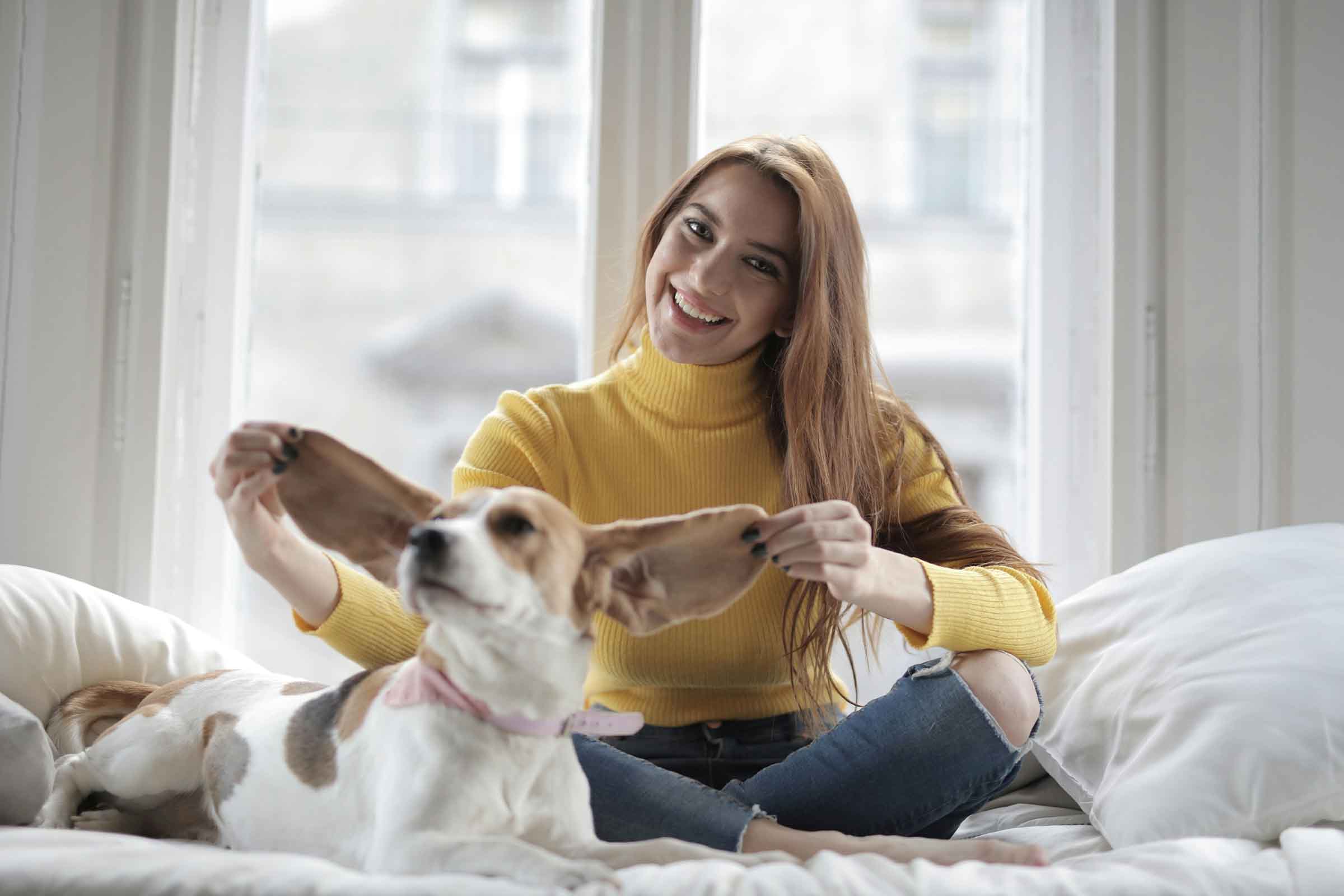 Femeie stând cu câinele pe pat și ținându-i urechile în sus