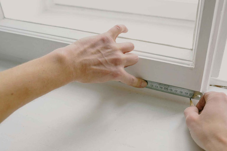 O persoană măsoară o ramă de fereastră albă cu o riglă metalică și un creion.