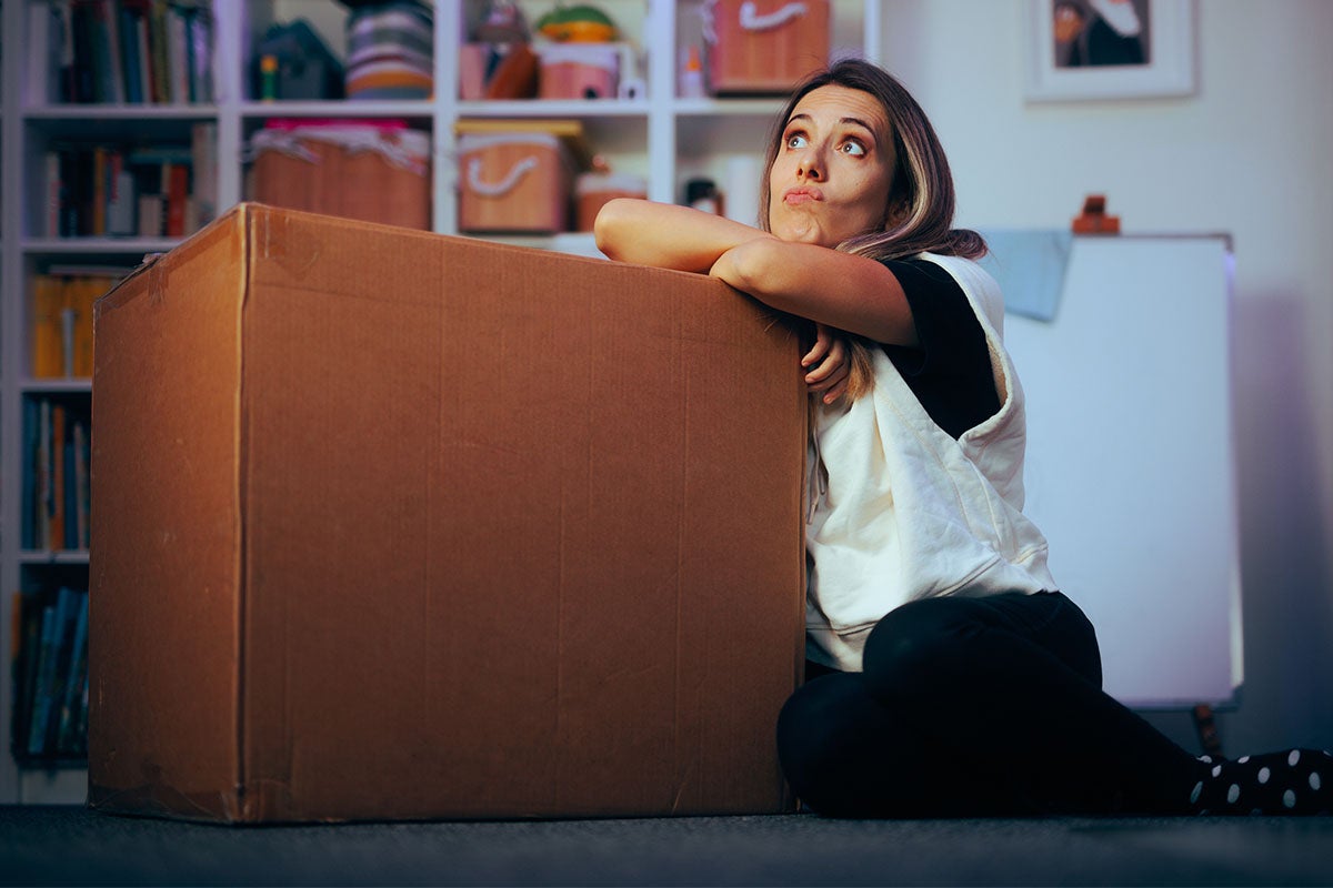 Femeie sprijinită de o cutie mare de carton în sufragerie în fața unui raft.