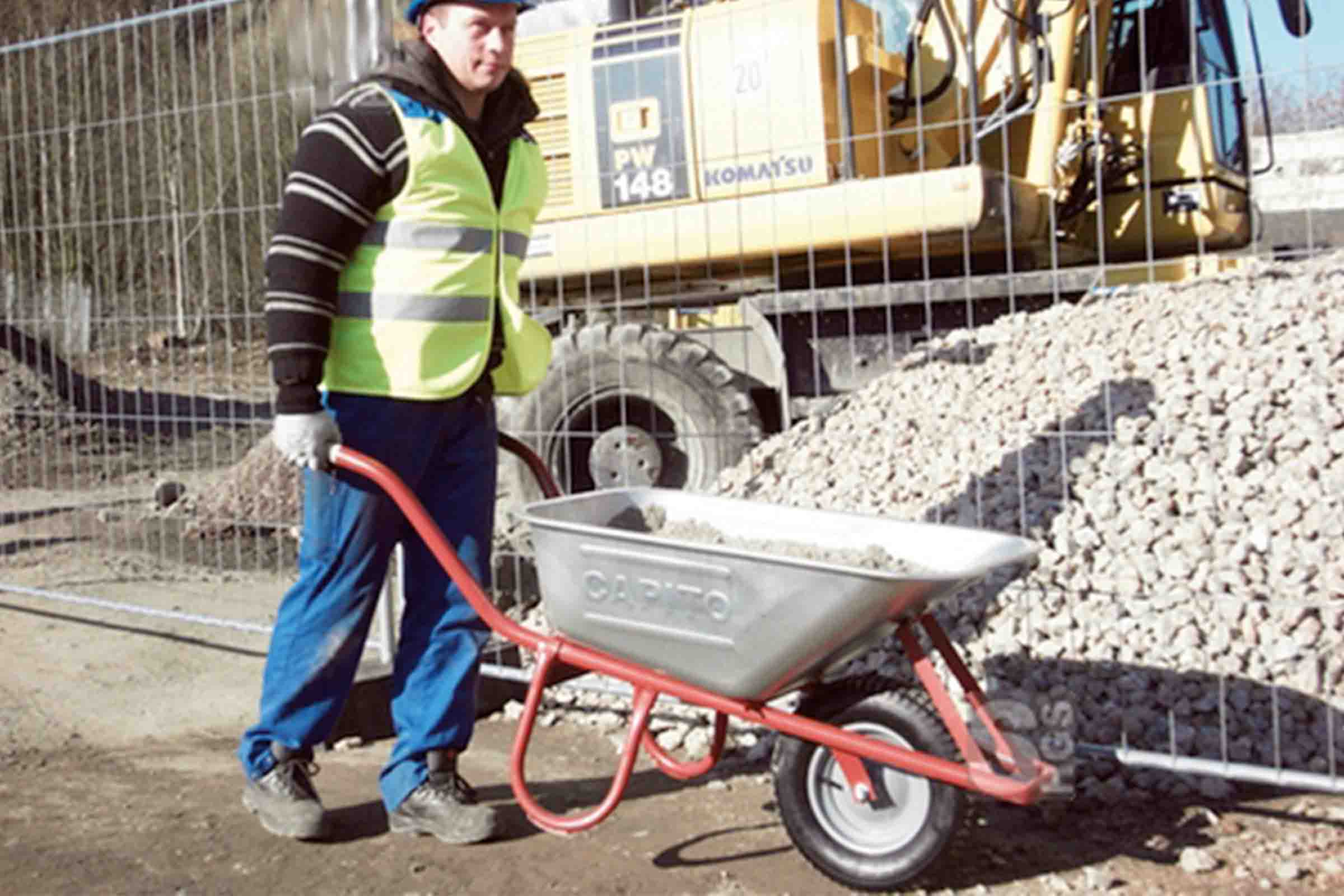Muncitor în construcții împinge roaba cu pietriș în fața unui excavator