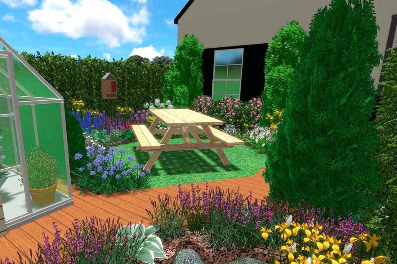 Grădină cu o masă de picnic din lemn pe gazon, înconjurată de straturi de flori, o terasă din lemn și o seră.
