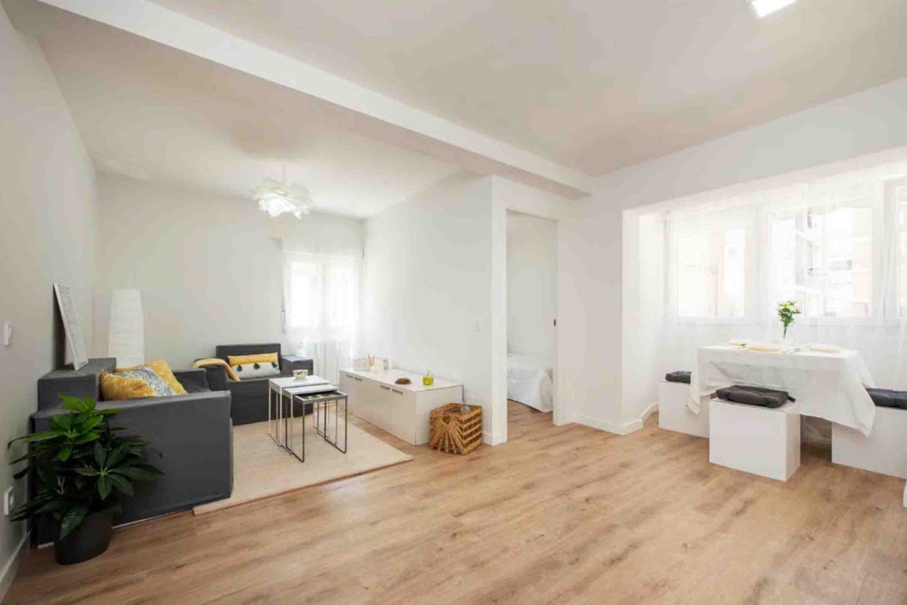 Cameră de zi luminoasă și spațioasă cu pereți albi, pardoseală aspect lemn, canapea gri închis și mobilier alb modern.