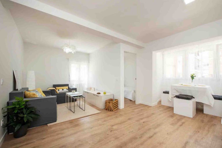 Cameră de zi luminoasă și spațioasă cu pereți albi, pardoseală aspect lemn, canapea gri închis și mobilier alb modern.