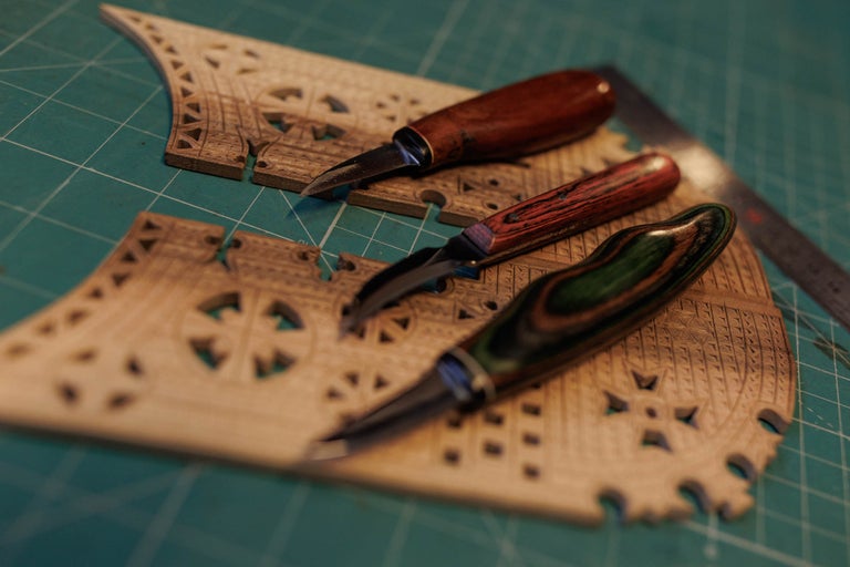 Trei cuțite de sculptat în lemn cu mânere din lemn așezate pe o bucată de lemn sculptată.