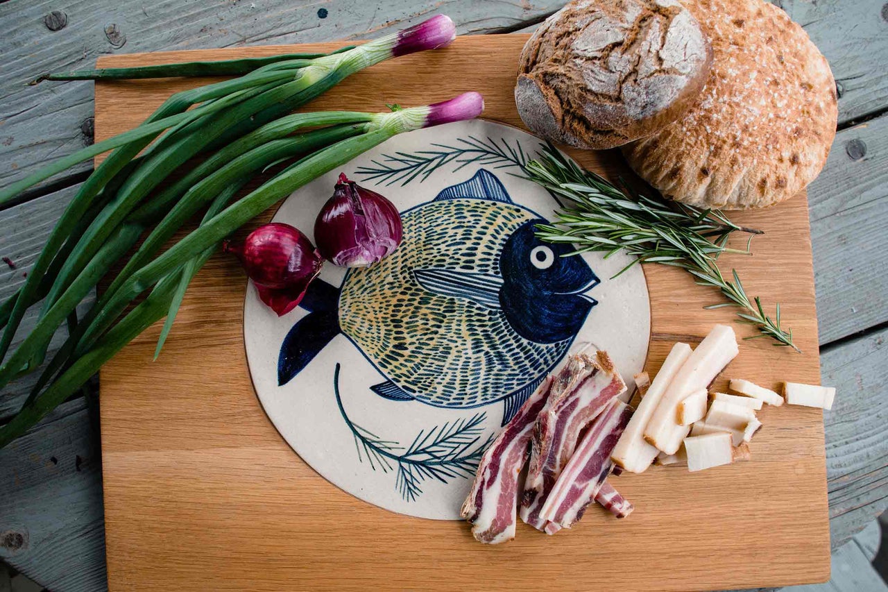 Placă de lemn cu ingrediente alimentare: pâine, ceapă, bacon și farfurie cu motiv de pește