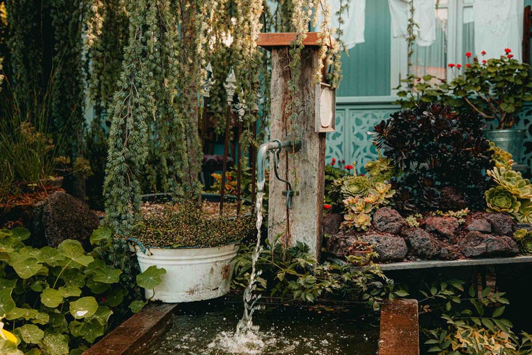 Fântână rustică de grădină cu stâlp de lemn și pipă în grădină cu plante, pietre și ghivece.