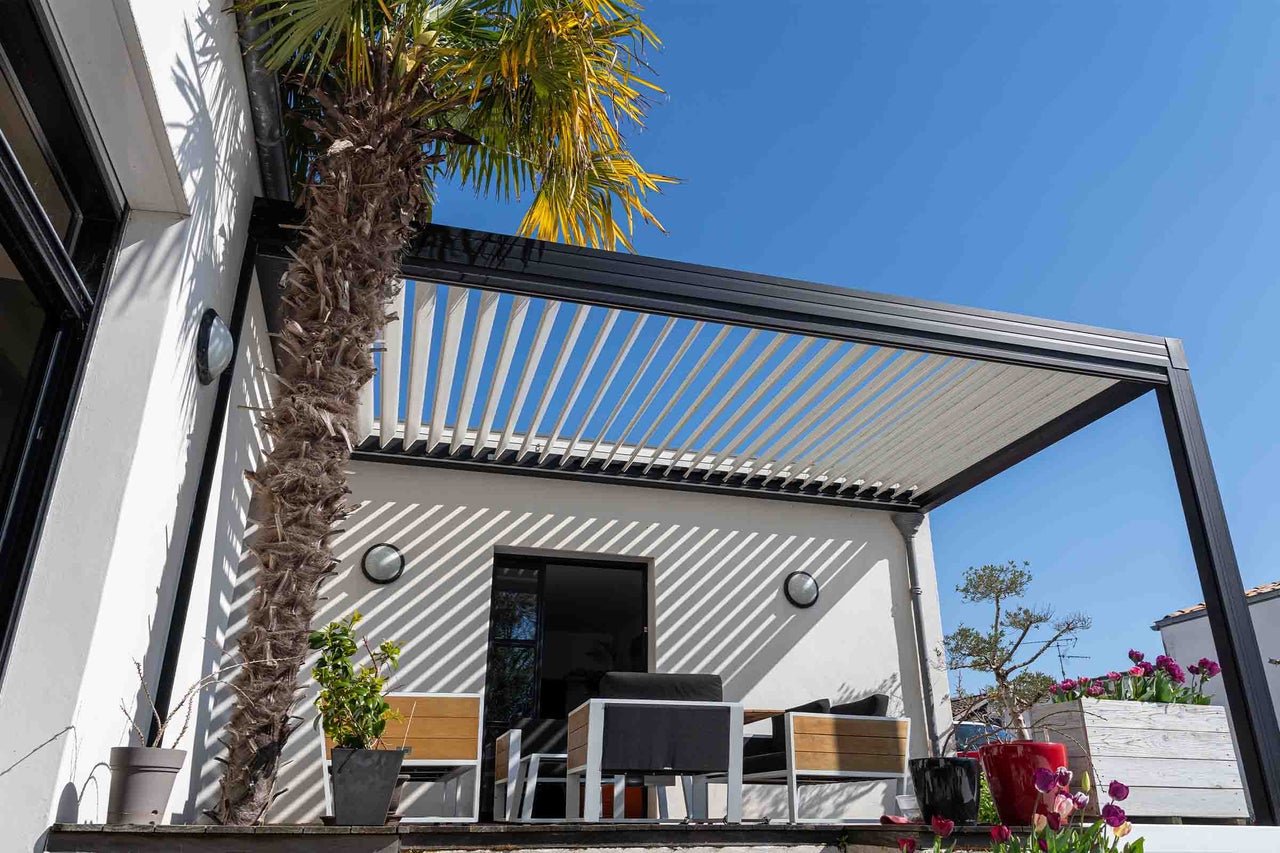 Pergolă modernă cu lamele antracit și lamele albe, fixată pe peretele casei, deasupra unei terase luminoase cu mobilier de grădină și palmier.