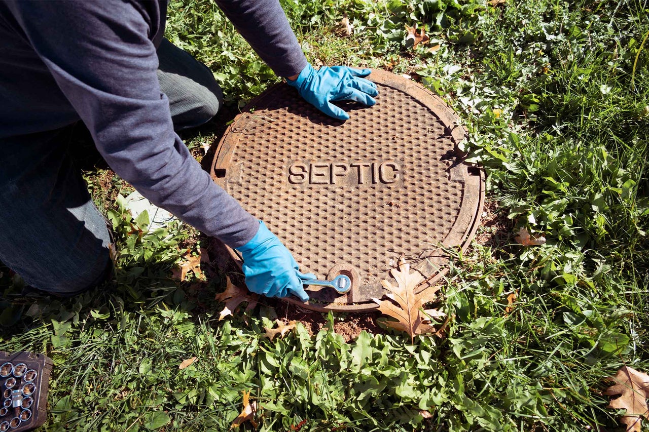 Persoană care deschide un capac metalic rotund de fosă septică cu inscripția Septic pe o suprafață de gazon.