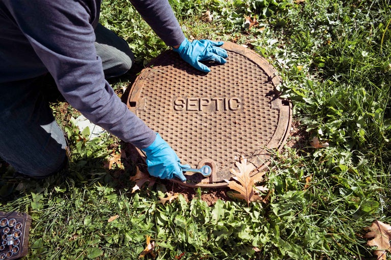 Persoană care deschide un capac metalic rotund de fosă septică cu inscripția Septic pe o suprafață de gazon.