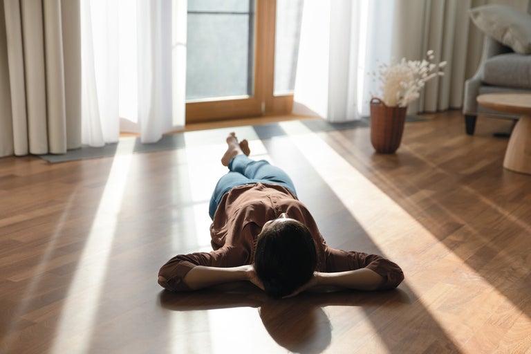 O persoană stă întinsă pe o pardoseală din parchet de lemn într-o cameră de zi luminoasă, cu lumina soarelui intrând prin ușa terasei.