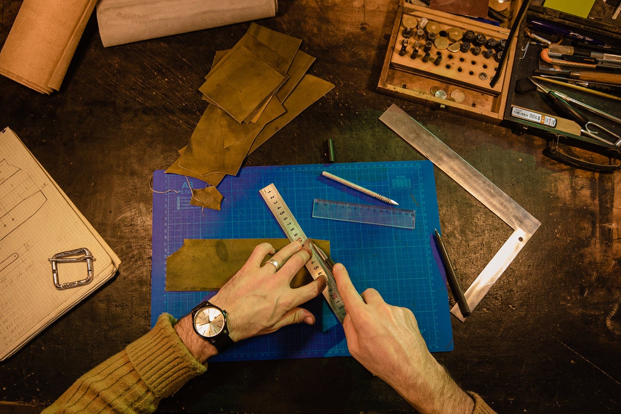 Scenă de atelier cu mâini tăind piele cu un cutter rotativ pe o placă de tăiat, înconjurate de unelte și materiale pentru prelucrarea pielii.