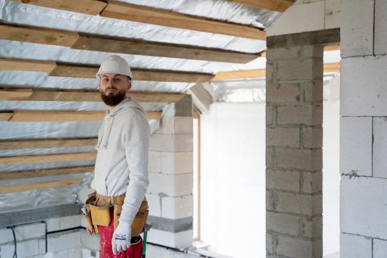 Muncitor cu cască de protecție albă și centură de scule pe un șantier în mansardă cu izolație și zidărie.