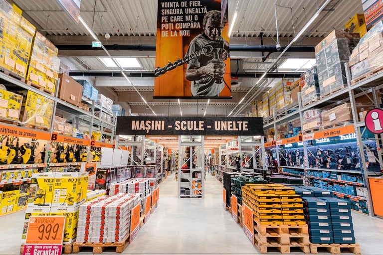 Vedere spre culoar într-un magazin Hornbach cu rafturi pline cu unelte și mașini
