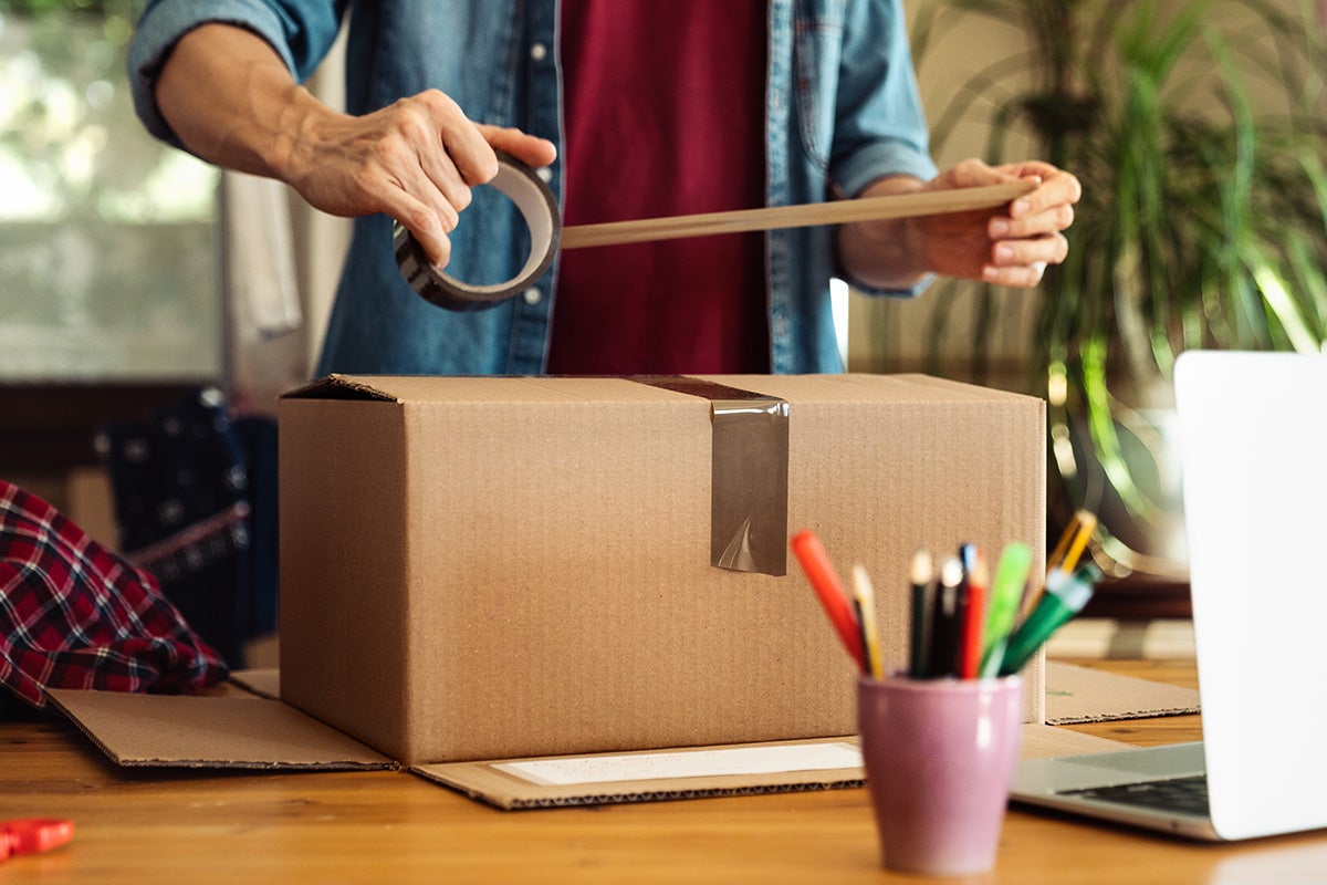 O persoană împachetează o cutie de carton cu bandă adezivă pe o masă.