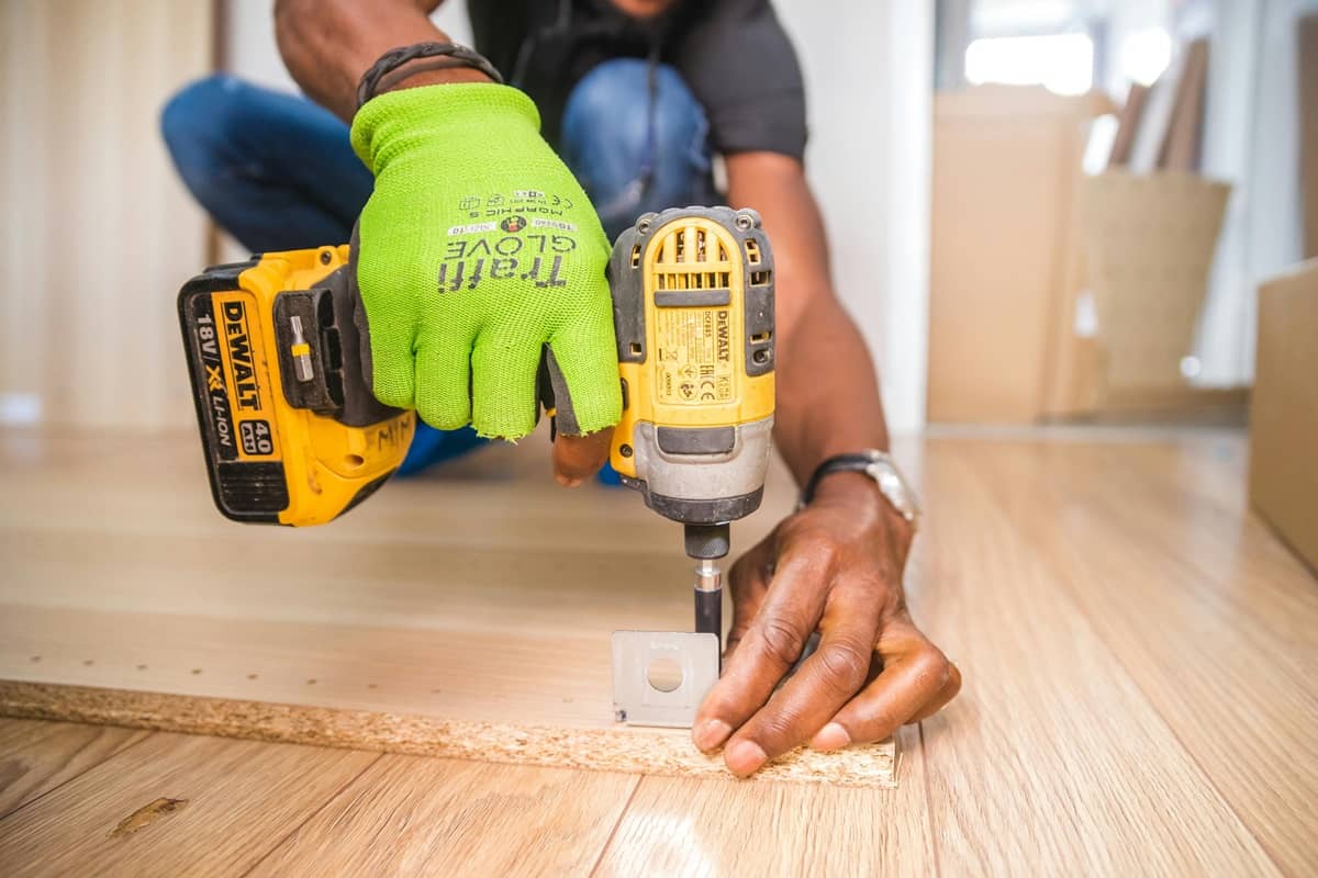 O persoană fixează o piesă de mobilier cu o șurubelniță electrică și poartă mănuși de lucru.