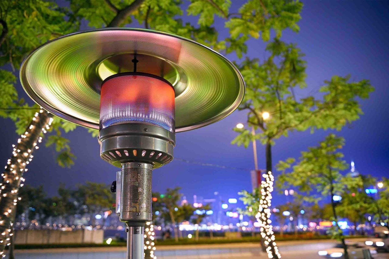 Încălzitor de terasă pe gaz într-un parc noaptea cu arzător iluminat și reflector metalic în fața copacilor cu ghirlande luminoase.