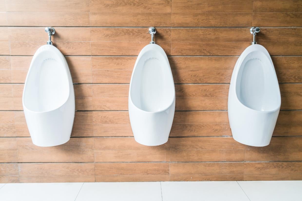 Trei pisoare suspendate din ceramică albă în fața unui perete cu plăci aspect lemn într-o baie modernă.